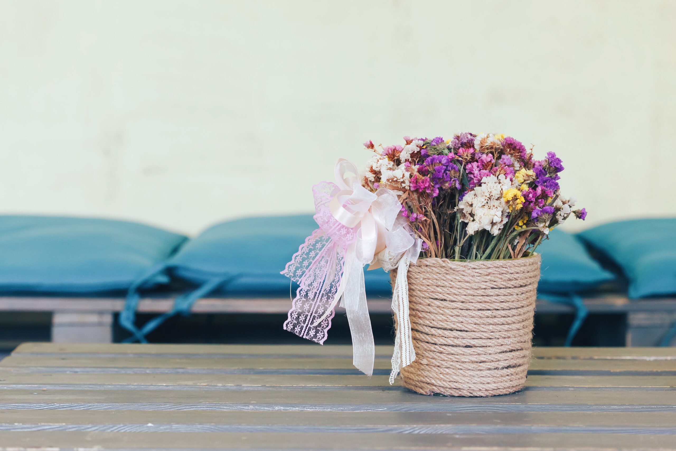 Doniczka z kolorowymi kwiatami owinięta jutowym sznurkiem, stojąca na drewnainym stole w pomieszczeniu