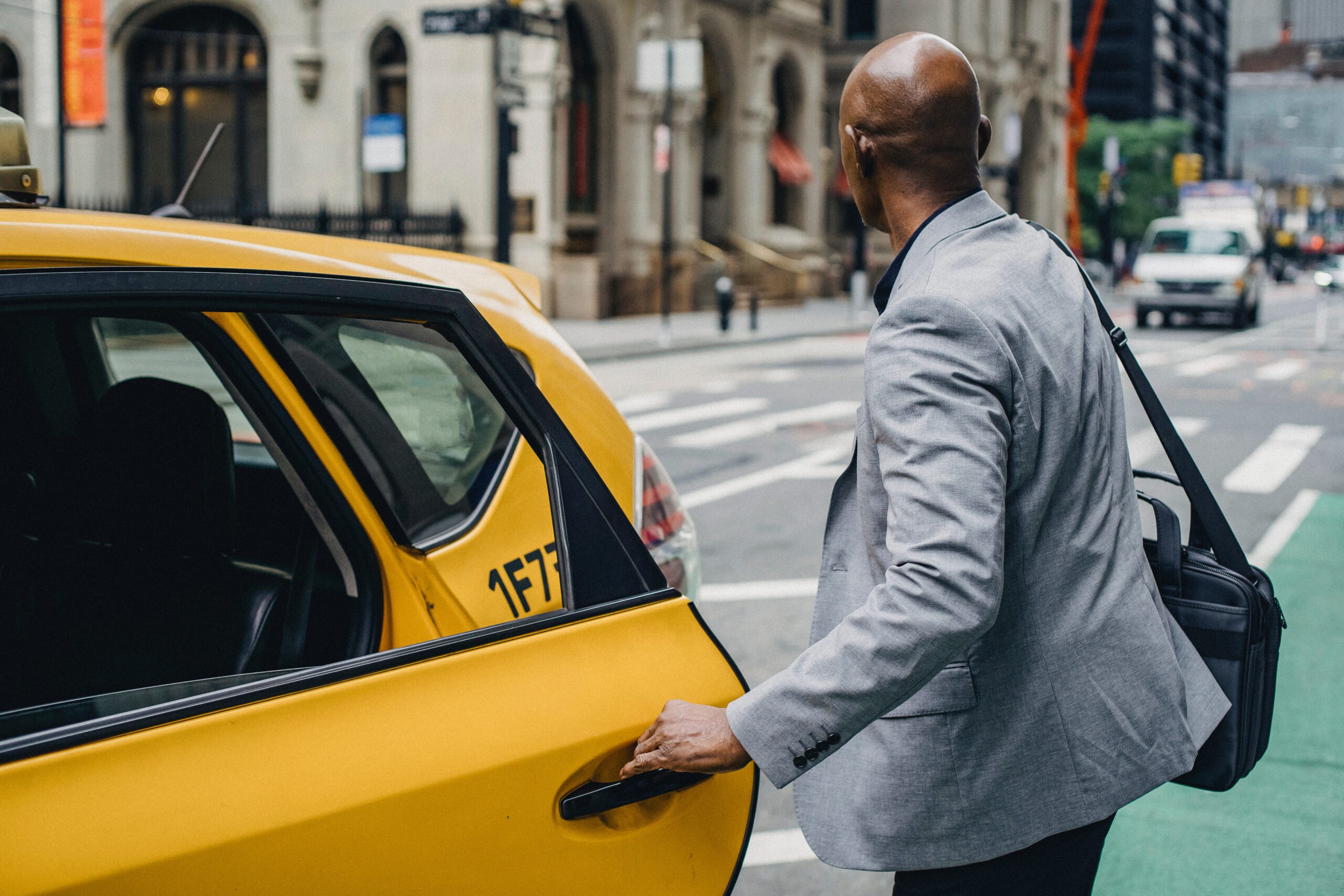 Mężczyzna ubrany w stylu smart casual z teczą na ramieniu, wsiadający do żółtej taksówki