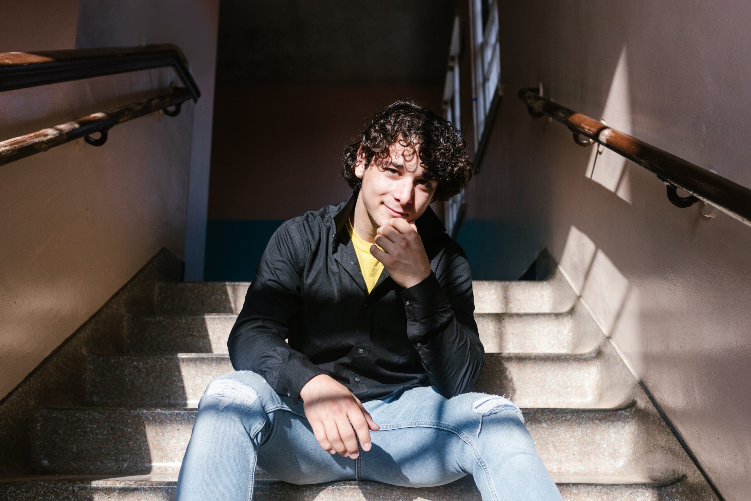 Usmiechający się młody mężczyzna, siedzący na schodach, ubrany w jeansy z dziurami i przetarciami na kolanach